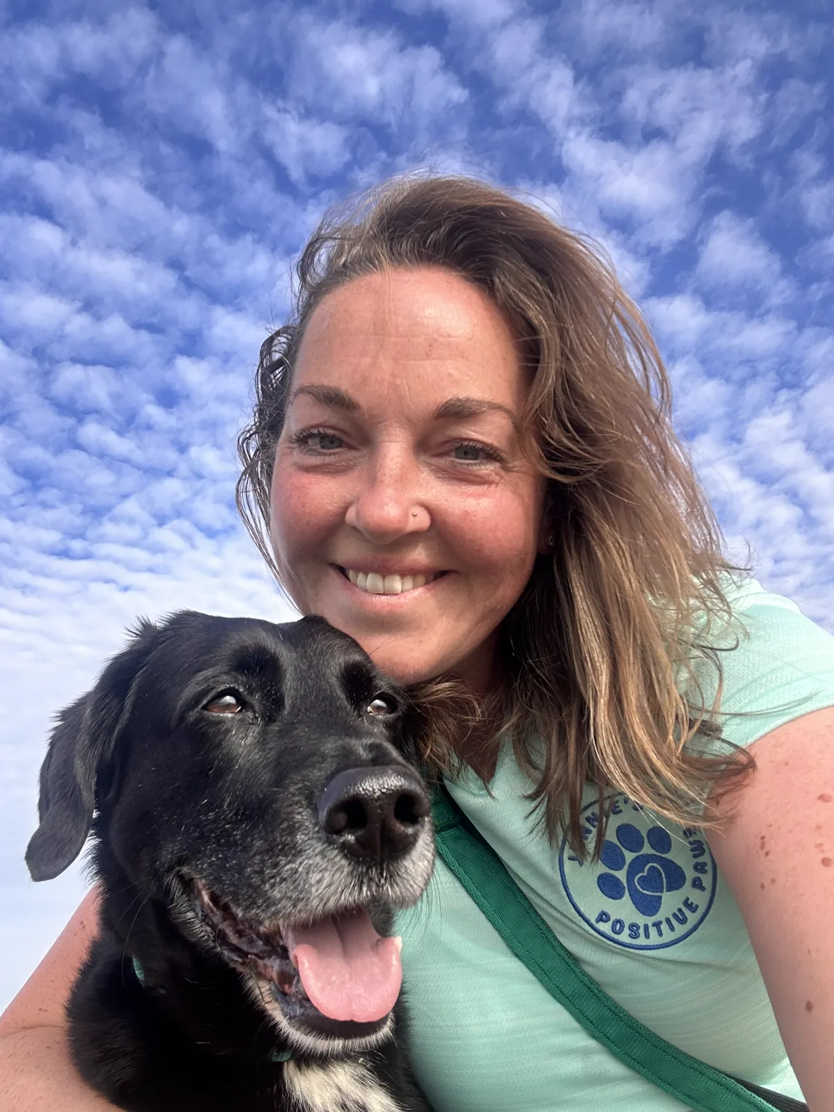 Jennie from Jennie's Positive Paws smiling with a black dog under a blue sky